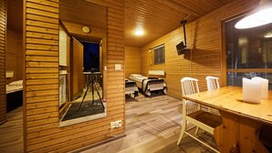 Een houten blokhut van binnen met een eettafel, stoelen, een bed en een raam met jaloezieën.