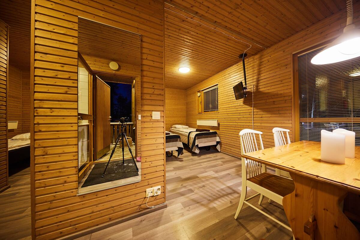 Interior de una cabaña de madera con una mesa de comedor, sillas, una cama y una ventana con persianas.