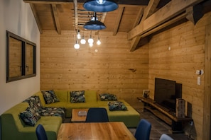 A cozy room with a wooden ceiling, a green sofa with patterned cushions, a wooden table, and a TV on a wooden stand.