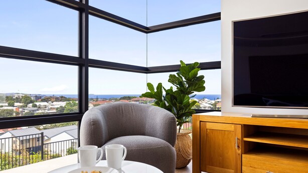 A modern room with a large window, a grey armchair, a white table with a plate of cookies and a cup, and a wooden cabinet with a TV.