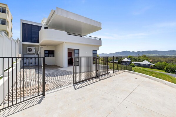 A modern two-story house with a balcony, a gated entrance, and a view of the surrounding landscape.