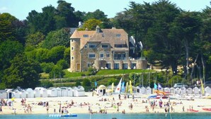 A large stone building with multiple windows and a tower, located near a beach with people and boats. - -GAÏA- MAISON FAMILIALE VUE MER ET JARDIN DOURVEIL (Névez)