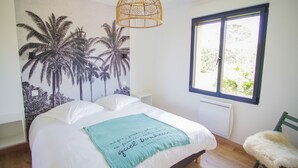 A bedroom with a bed, a wall mural of palm trees, a window, and a green chair. - -GAÏA- MAISON FAMILIALE VUE MER ET JARDIN DOURVEIL (Névez)