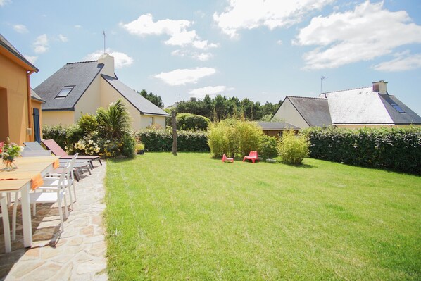 A well-maintained backyard with a stone pathway leading to a table and chairs, surrounded by lush greenery and a clear sky. - -GAÏA- MAISON FAMILIALE VUE MER ET JARDIN DOURVEIL (Névez)