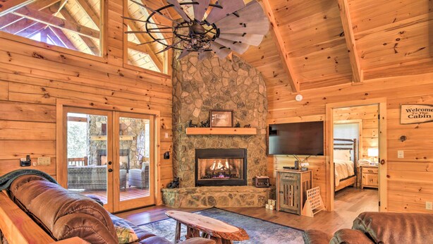 A cozy cabin interior with a stone fireplace, leather seating, and a ceiling fan. - Private Cabin w/ Furnished Porch on < 3 Acres! (Mineral Bluff)
