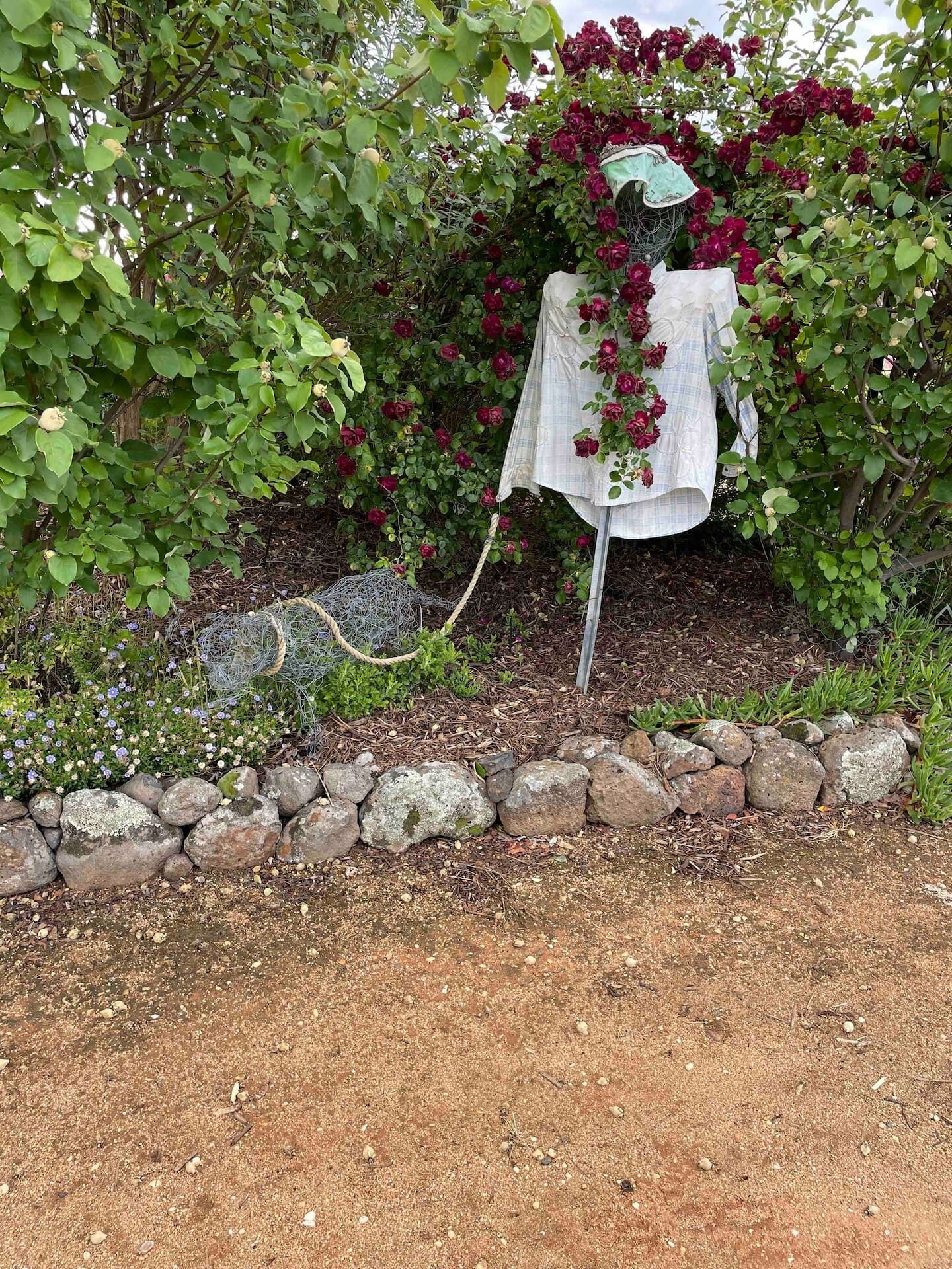 Ein Garten mit einer Steinmauer, einem Baum mit grünen Blättern und roten Blüten sowie einem weißen Tuch, das über einen Metallpfosten drapiert ist.
