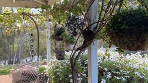Un espacio al aire libre techado con un sillón colgante, macetas suspendidas y un jardín con flores blancas.