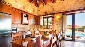 A kitchen with a stone wall, wooden cabinets, a stainless steel refrigerator, and a dining area with a glass table and chairs. - Traditional Holiday Villa with Panoramic View (Chania)