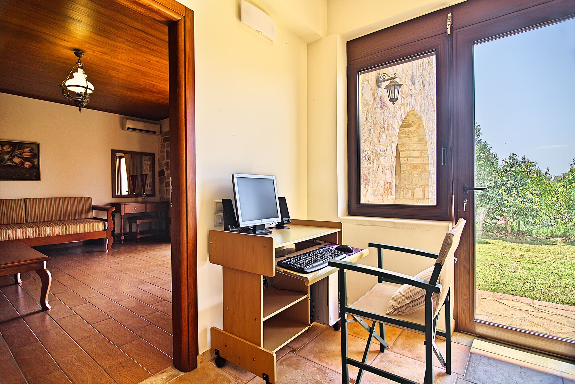 Een kamer met een houten bureau, computer en stoel, een bank en een groot raam met uitzicht op groen.