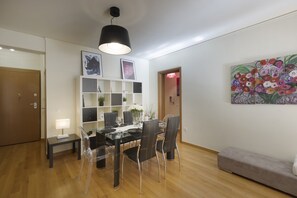 A modern dining area with a glass table, black chairs, a white shelving unit with decorative items, and a colorful floral painting on the wall. - MisaraliotouA3 · 3BR Prime APT a breath away from (Athens)
