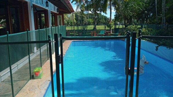 A swimming pool with a green fence, surrounded by a garden and a building with a wooden facade. - Villa Casa Nostra by KlabHouse 5BDRM w/Pool and Chef (La Romana)