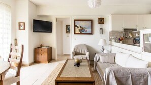 A modern living room with a sofa, coffee table, and TV. There is a dining area with a table and chairs. The room has a neutral color palette and a large window. - Compact 3 room apartment in Mall Athens. (Marousi)