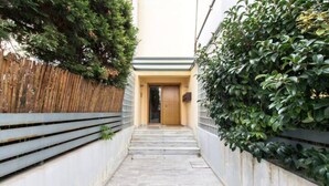 A narrow, tiled pathway leading to a wooden door, flanked by a bamboo fence and dense green foliage. - Compact 3 room apartment in Mall Athens. (Marousi)