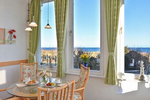 A dining area with a glass table, chairs with floral upholstery, and a view of the sea through green curtains. - Meerblick 05 House - with a direct view of the sea (Ahlbeck (Seebad))