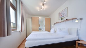 Una habitación de hotel moderna con una cama grande, suelo de madera, un espejo, un estante en la pared con un jarrón y una lámpara en el techo.
