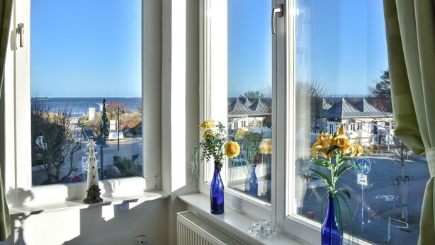 Una ventana con vista al mar y edificios, un radiador blanco y dos jarrones con flores.
