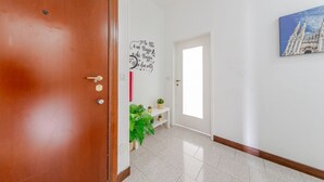 A hallway with a wooden door, a white wall with a decorative sign, a small shelf with plants, and a framed picture. - GuestHost - Bonola M1 Underground Bright & Functional Apt x4 (Milano)