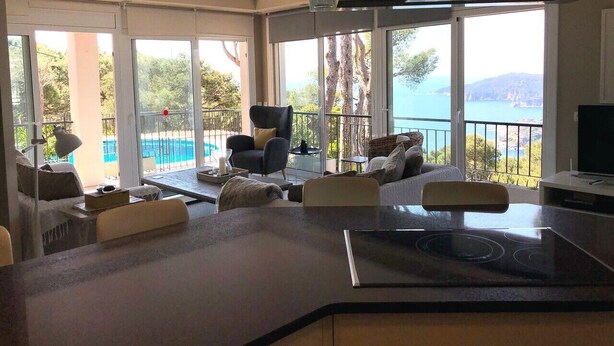 Un salon moderne avec un coin cuisine, de grandes fenêtres offrant une vue sur la mer et un balcon aménagé avec du mobilier d’extérieur.