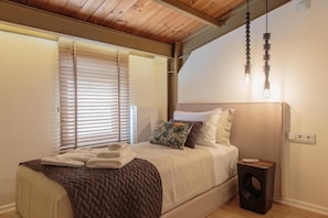 A bedroom with a bed, a bedside table, a window with blinds, and a hanging light fixture.