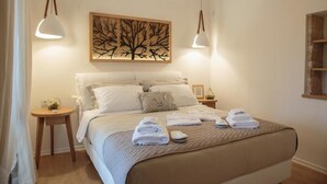 A neatly made bed with a quilted cover, two bedside tables with lamps, and a wall-mounted artwork of a tree. - Unique vacation in traditional Cretan village in the mountains with family (Zaros)