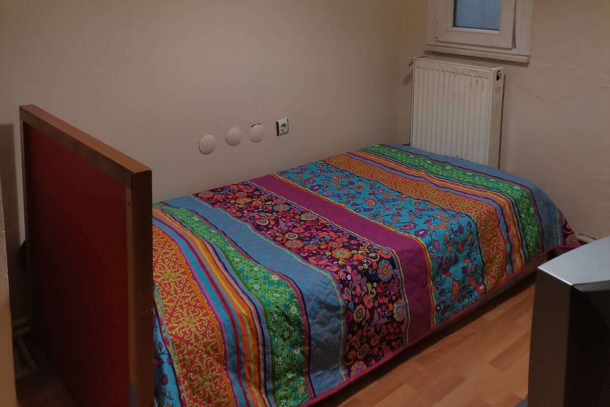 Quarto pequeno com uma cama de solteiro, um edredom colorido, uma janela com vista, um radiador e um quadro na parede.