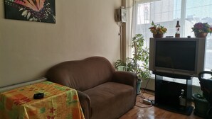 A living room with a brown sofa, a table with a floral tablecloth, a television on a stand, and several potted plants. - Marina Pansiyon (Silivri)