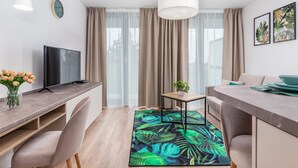 A modern living room with a flat-screen TV, a dining area with a table and chairs, a coffee table with a plant, and a large window with curtains. - Air-conditioned Studio 50 m from the Beach | Parking | Gym (Mielno)