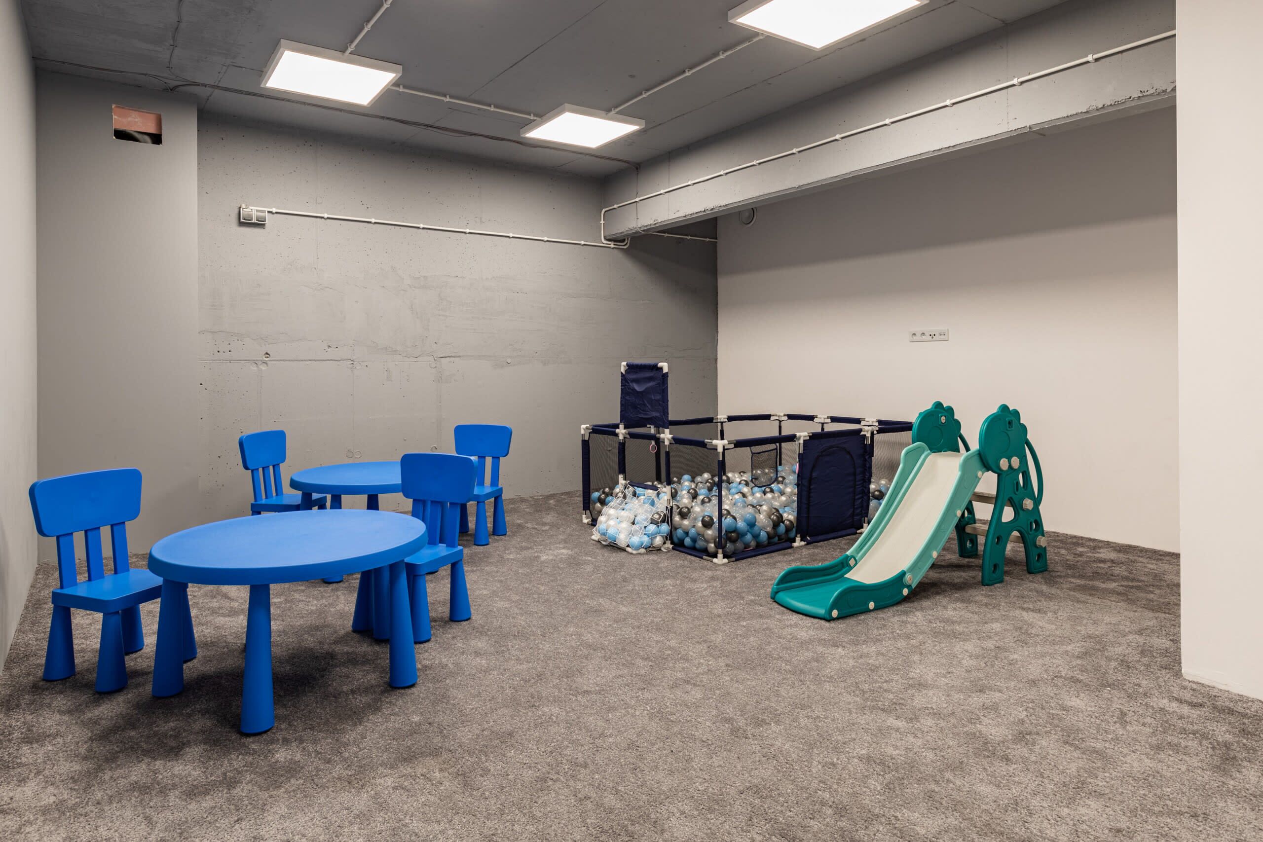 Ein Spielbereich mit blauen Stühlen und Tischen, einem Spielbereich mit Bällen und einer grünen Rutsche.