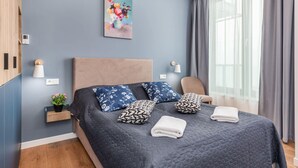 A modern bedroom with a large bed, a bedside table with a lamp, a small plant, and a painting on the wall.