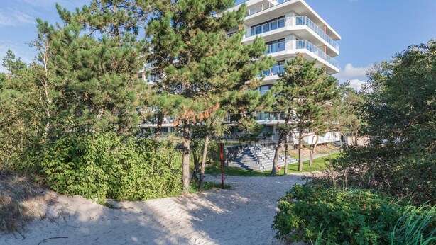 A modern multi-story building with balconies, surrounded by dense greenery and a sandy area.
