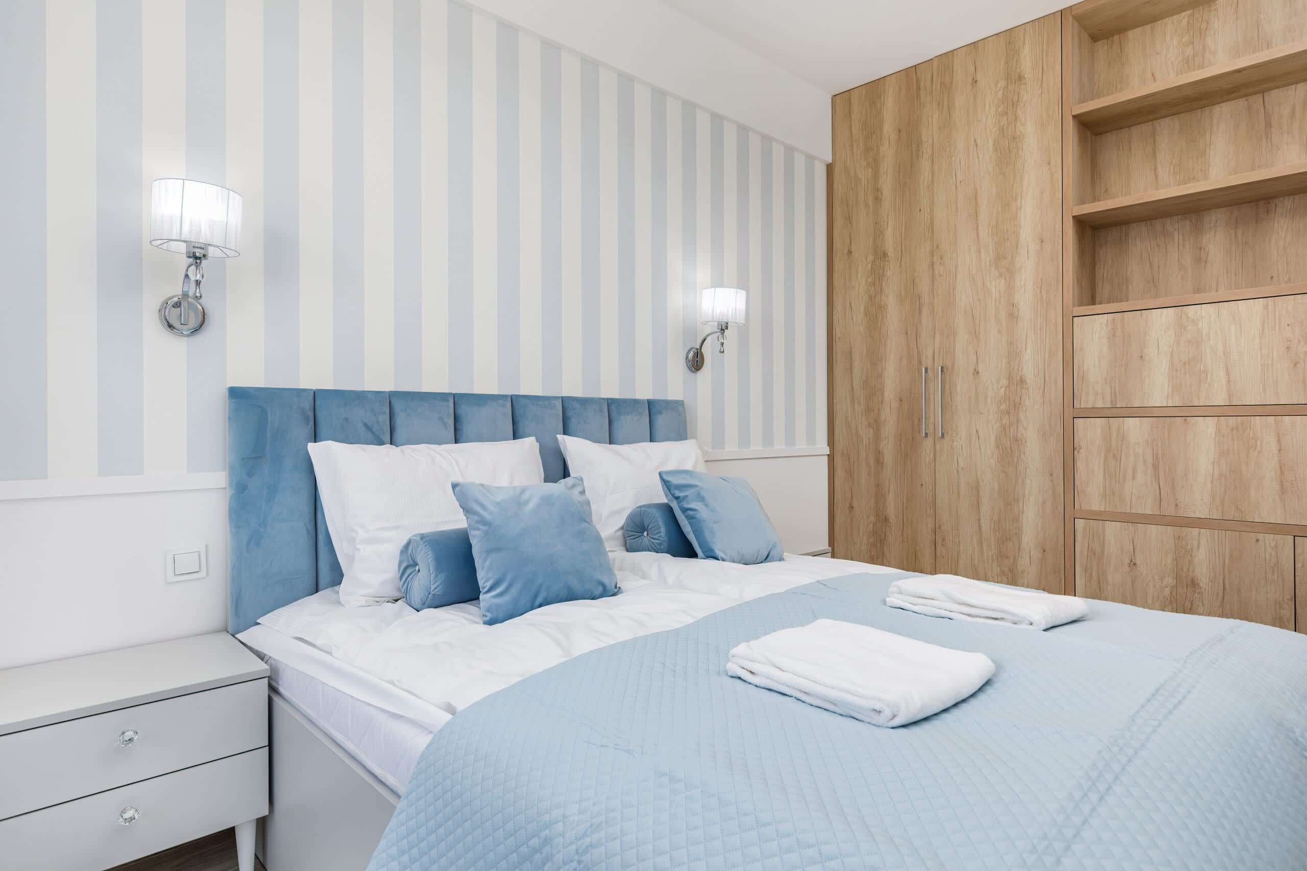 A bedroom with a striped wall, a wooden wardrobe, a bed with blue bedding, and two wall-mounted lamps.