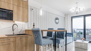 A modern kitchen with a microwave, sink, and wooden cabinets. There is a dining area with blue upholstered chairs and a white coffee table. A large window offers a view of the city.
