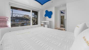 A modern bedroom with a large bed, a window with a view of a building, a blue accent wall, and a small bathroom visible through an open door.