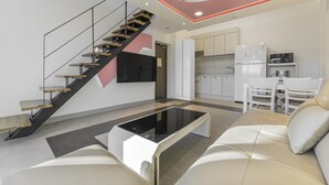 Una sala de estar moderna con una escalera, un televisor de pantalla plana, una zona de cocina con armarios blancos y una mesa de comedor con sillas.