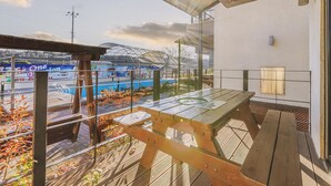 Un patio al aire libre con mesa y sillas de madera, baranda de vidrio y vista a una piscina y un edificio con estructura en forma de cúpula.