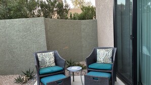 Twee rieten stoelen met teal kussens en bedrukte kussens, een klein rond tafeltje, en een betonnen terras met een textuurwand en planten.