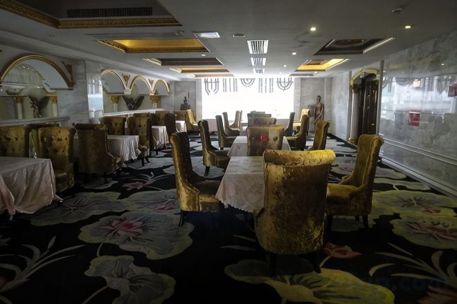 Un salón comedor de hotel con mesas redondas, sillas doradas y una alfombra con motivos florales.