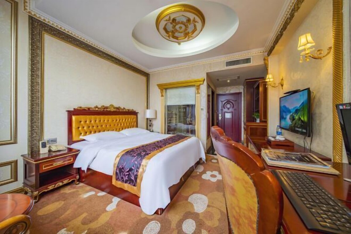 A hotel room with a large bed, a desk with a computer, a television, and a door leading to another room.