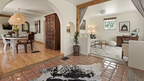 Un salon spacieux comprenant un coin repas, des poutres apparentes, un grand lustre et un tapis rustique.