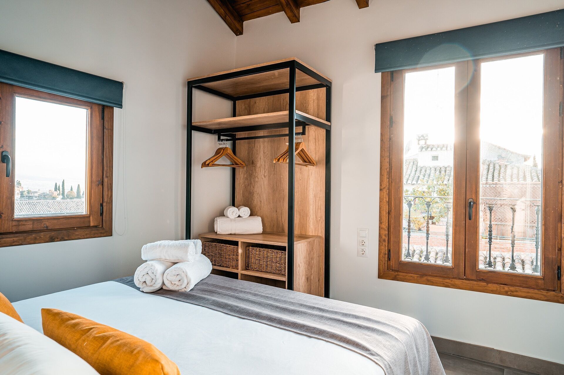Un dormitorio con cama, un armario de madera con estantes, una ventana con vista a edificios y una barandilla de balcón.