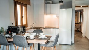 A modern kitchen with white cabinets, a built-in oven, and a refrigerator. There is a dining table with chairs and a coffee machine. - Description (Granada)