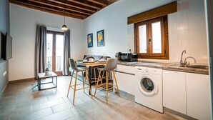 Una cuina compacta amb rentadora, zona de menjador amb cadires i televisió penjada a la paret.