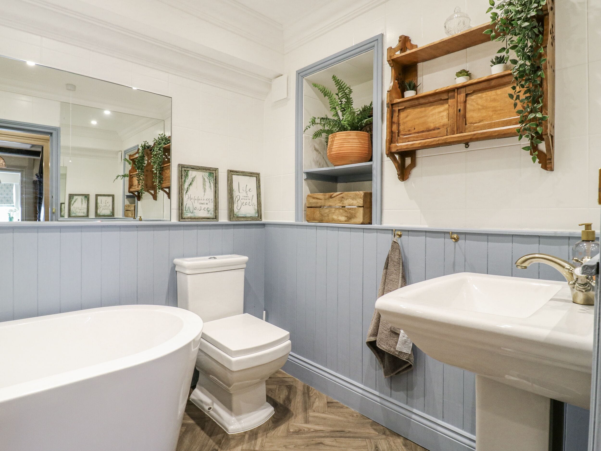 Et badeværelse med et hvidt badekar, toilet og vask. Der er et spejl over vasken, og en hylde med planter og pynteting.