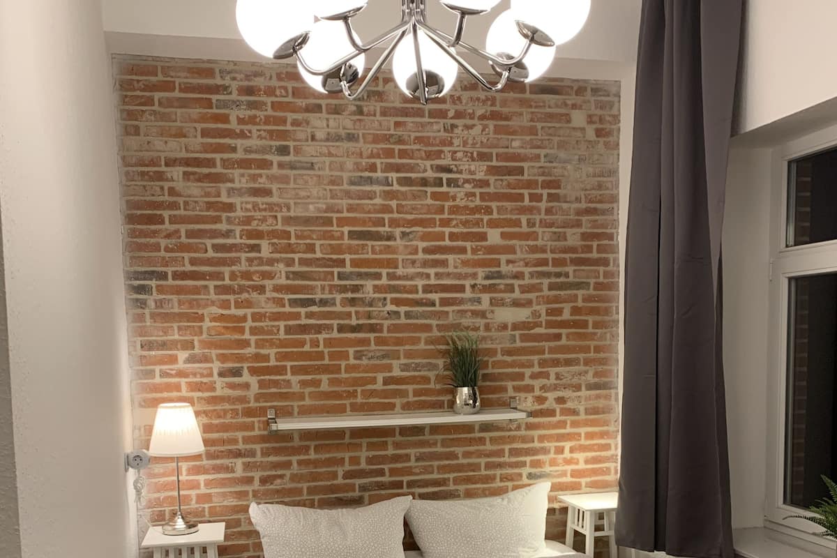 Un dormitorio con pared de ladrillo, una araña, una mesita de noche con lámpara, una ventana con cortinas y una cama con almohadas y una colcha.