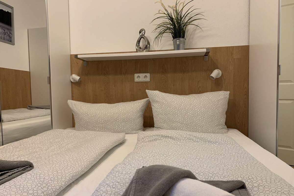 Una cama bien hecha con ropa de cama estampada y una manta gris, una pequeña planta en maceta en un estante encima y un cabecero de madera.