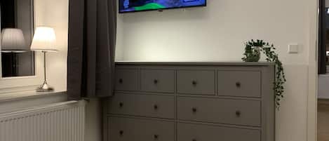 Habitación de hotel con una cama, un aparador gris, un televisor en la pared, una ventana con cortinas, una lámpara y una planta.