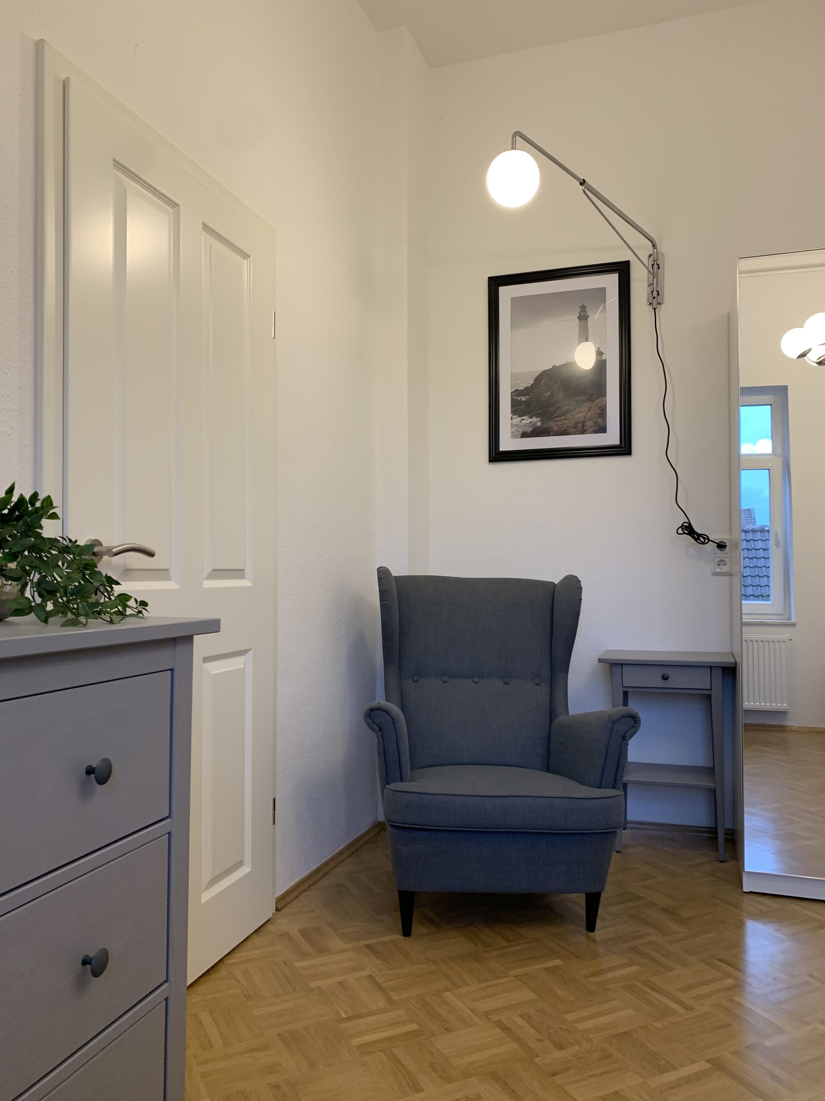 Una habitación con un sillón tapizado gris, un escritorio pequeño, un aparador blanco con cajones, un cuadro en la pared y una lámpara.