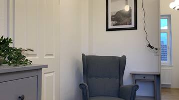 Una habitación con un sillón tapizado gris, un escritorio pequeño, un aparador blanco con cajones, un cuadro en la pared y una lámpara.