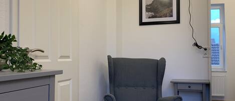 Une pièce comprenant un fauteuil à revêtement gris, un petit bureau, une commode blanche avec des tiroirs, un tableau encadré accroché au mur et une lampe.