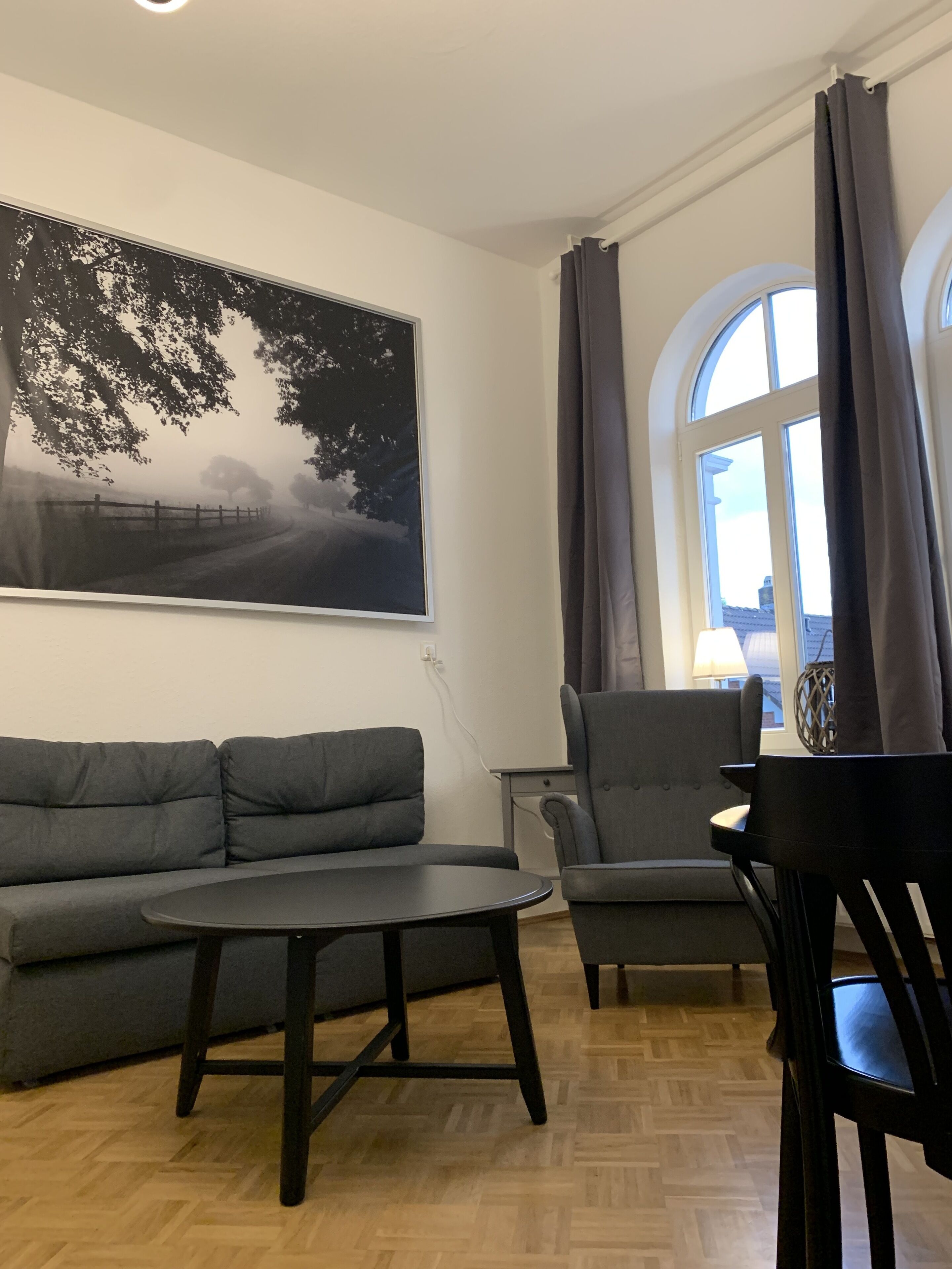 Una sala de estar con un sofá, una silla, una mesa, un cuadro y una ventana con cortinas.